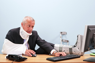 photo of a mature businessman with multiple injuries sitting at his desk struggling to work